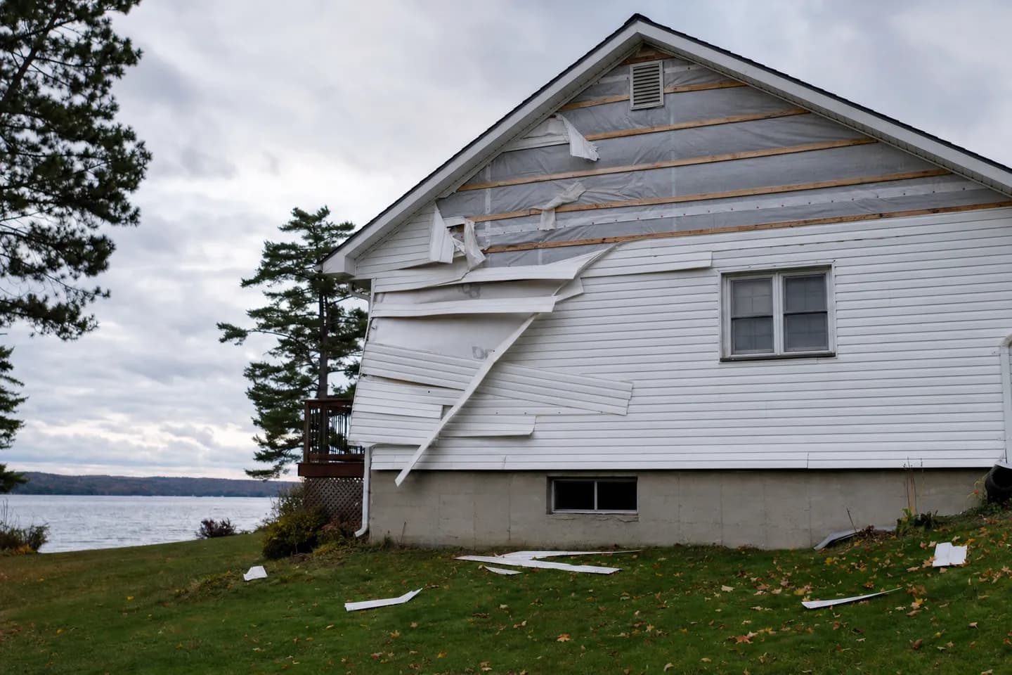 White vinyl siding torn off the gable of a lakeside Ontario house after a windstorm, with broken panels lying on the lawn