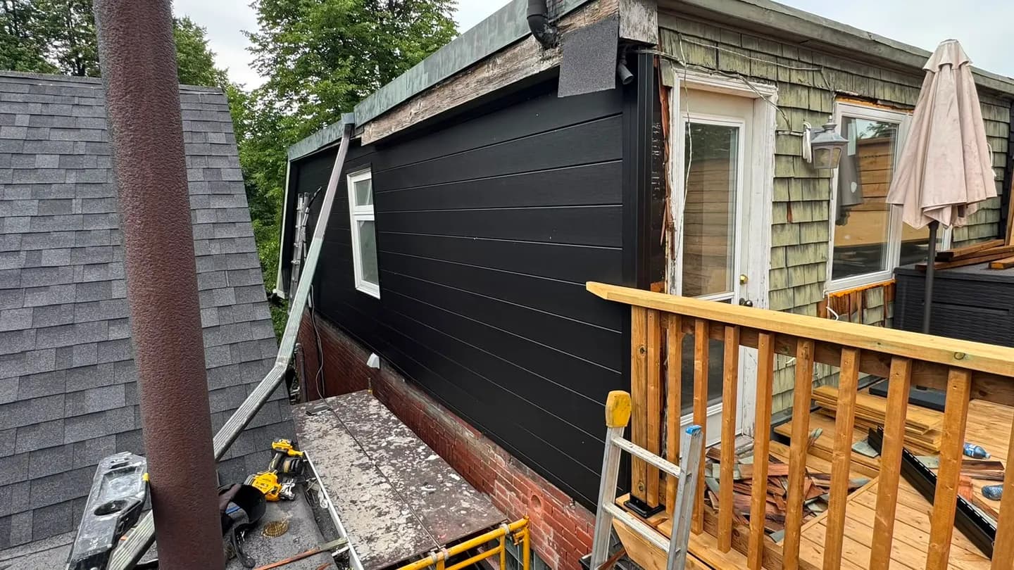 Side wall of the upper storey finished in black siding above the old brick, with a ladder and tools on the lower roof