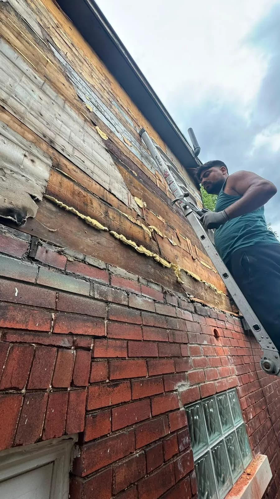 Old cladding layers stripped back above the brick, with a line of foam at the joint and a crew member on a ladder