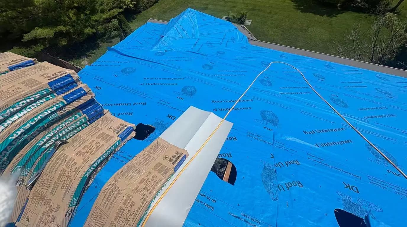 Blue synthetic roof underlayment laid over the new deck, with white drip edge along the rake and shingle bundles staged at the ridge
